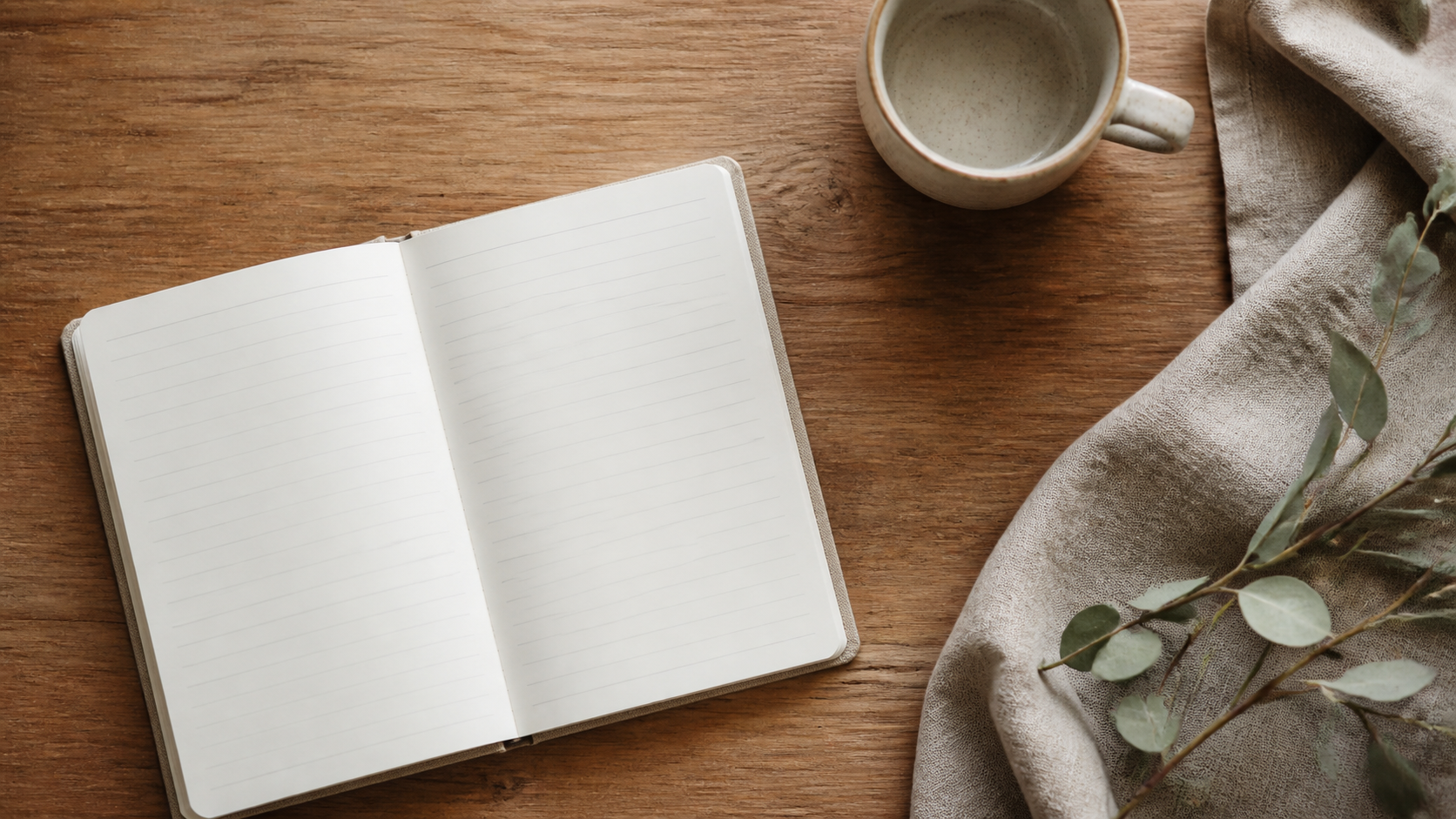 A blank notebook, ceramic cup, eucalyptus leaves and woven linen cloth on a warm timber desk.